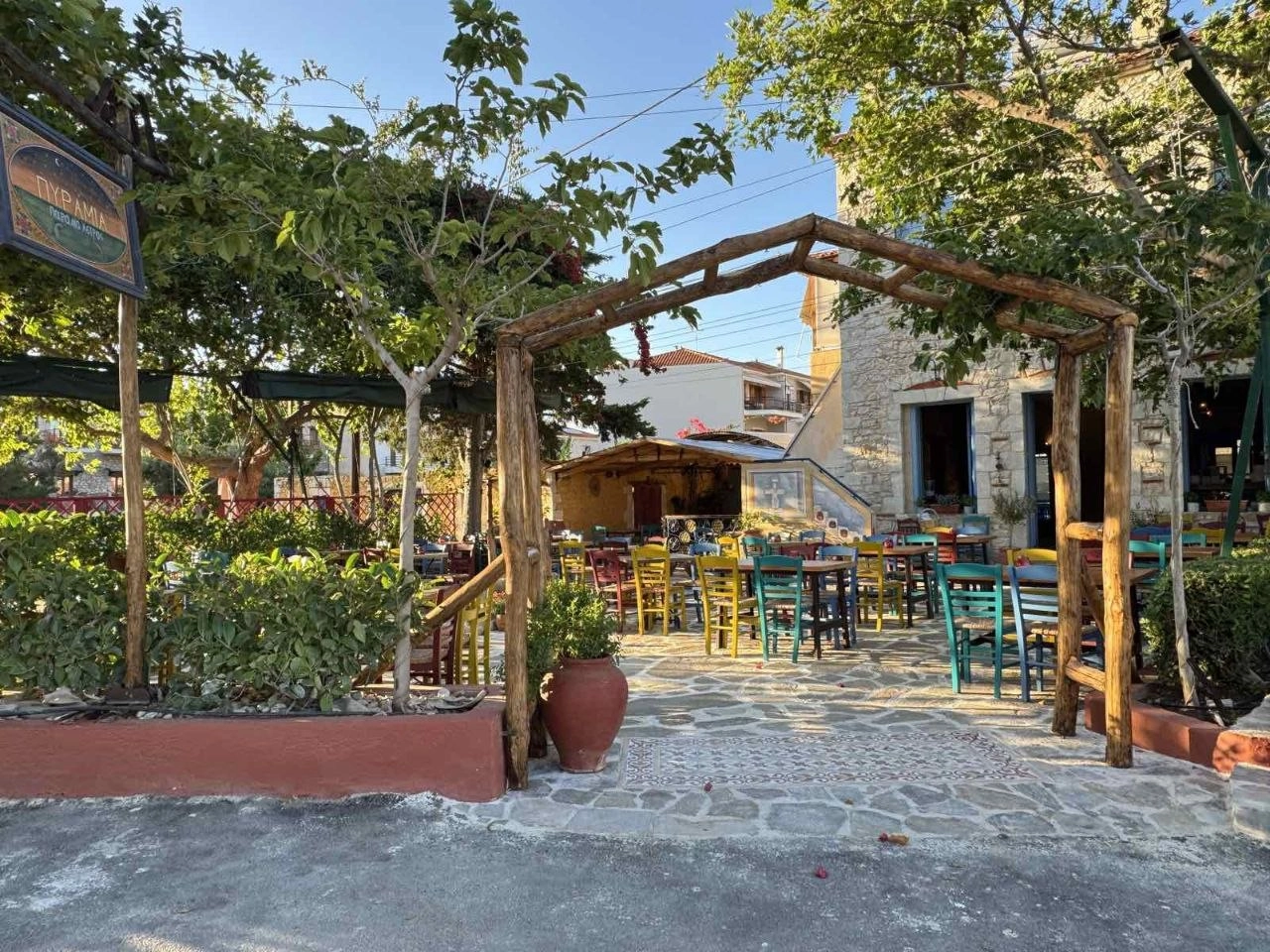 Η αυλή του Πυράμια την ημέρα με την πινακίδα,τραπέζια και δέντρα.